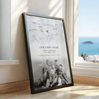 A framed personalised map print leaning against a window on a wooden floor, showing a faint map background, the text 'Our First Home' with names and date, and a black-and-white photo of a family at the bottom. A real interior setting and ocean view are visible through the window.