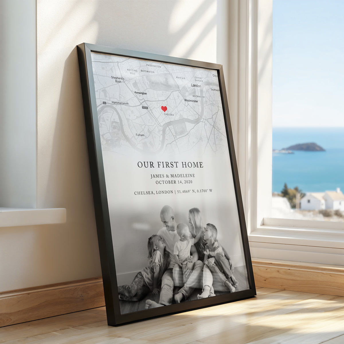 A framed personalised map print leaning against a window on a wooden floor, showing a faint map background, the text 'Our First Home' with names and date, and a black-and-white photo of a family at the bottom. A real interior setting and ocean view are visible through the window.