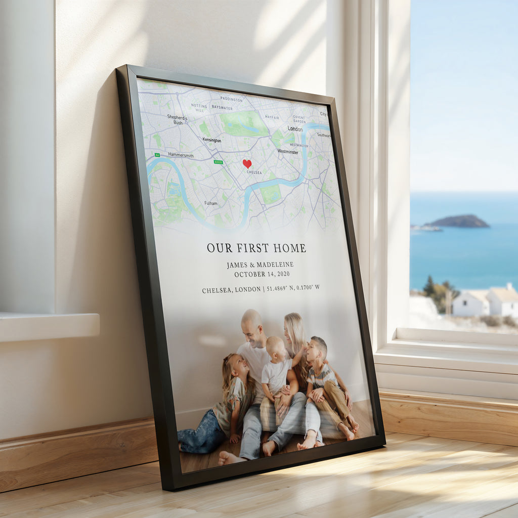 A framed personalised map print leaning against a window in a sunlit room. The print shows a map of Chelsea, London with a red heart marker and the text 'Our First Home' plus custom names and date with a photo of a family on the bottom.