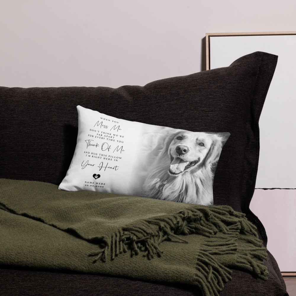 A rectangular personalised pet memorial throw pillow with a black and white photo of a dog and printed remembrance text, resting on a black sofa next to an olive green throw blanket.