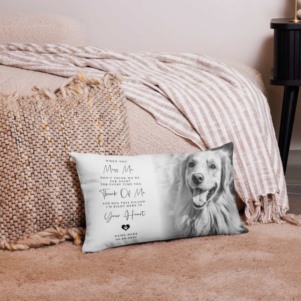 A rectangular personalised pet memorial pillow with a grayscale photo of a golden retriever and sentimental text, styled on a pink rug in front of a bed with a striped blanket.