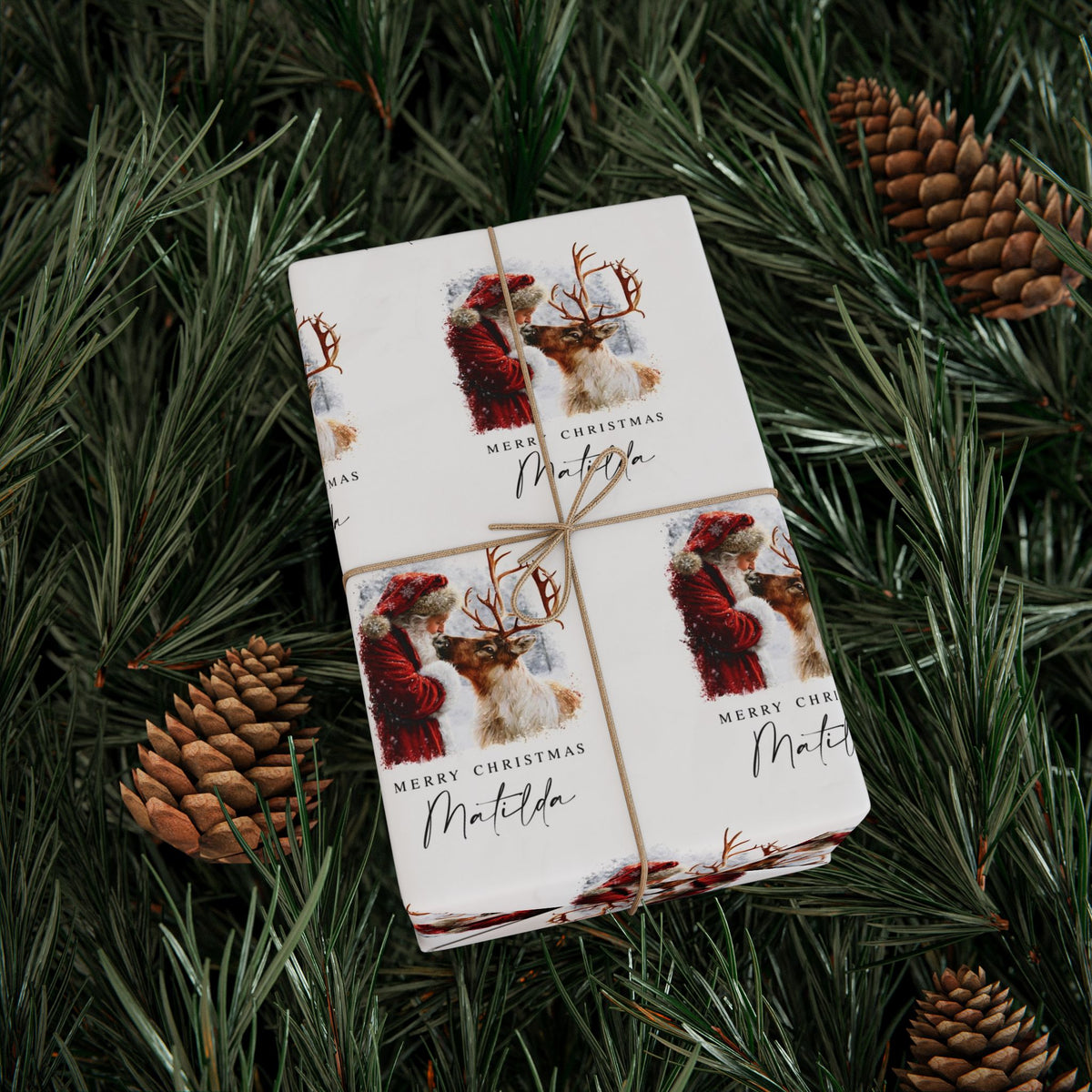A gift box wrapped in twine, displayed on a bed of green pine needles with pinecones. The wrapping paper features festive illustrations of Santa Claus and reindeer with the text 'MERRY CHRISTMAS Matilda'.