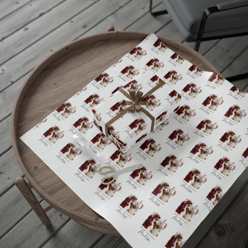 A gift wrapped box and a roll of matching wrapping paper displayed on a round wooden tray. The wrapping paper has a repeating print of a red and gold Santa Claus and Reindeer illustration with the name 'Matilda' written below each figure, and the box is tied with a brown ribbon.