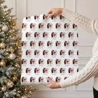 A person in a cream cable knit sweater holds up a large sheet of wrapping paper featuring a repeating pattern of vintage Santa Claus illustrations and the name 'Matilda.' A decorated Christmas tree with warm lights is visible in the background.