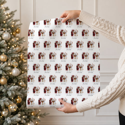 A person in a cream cable knit sweater holds up a large sheet of wrapping paper featuring a repeating pattern of vintage Santa Claus illustrations and the name 'Matilda.' A decorated Christmas tree with warm lights is visible in the background.