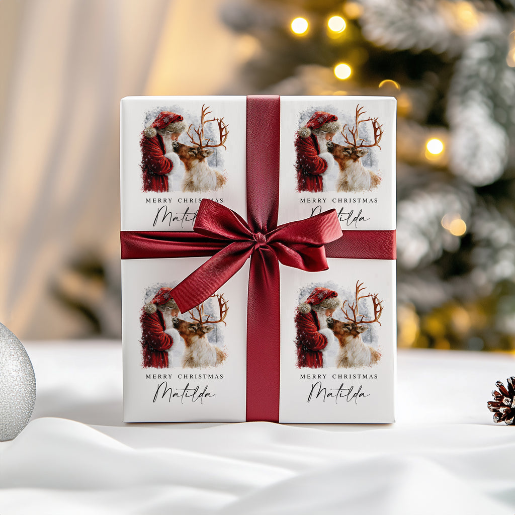 A square white gift box wrapped with a wide dark red ribbon and bow, printed with four Christmas illustrations of Santa and reindeer and the words 'Merry Christmas Matilda' The box is photographed on a white surface with a softly blurred festive background and warm lights.