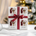 A square white gift box wrapped with a wide dark red ribbon and bow, printed with four Christmas illustrations of Santa and reindeer and the words 'Merry Christmas Matilda' The box is photographed on a white surface with a softly blurred festive background and warm lights.