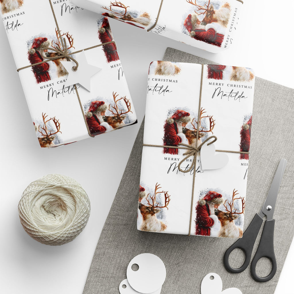 Three wrapped gift boxes with matching Christmas themed wrapping paper are arranged on a light grey surface with twine, a ball of string, scissors, and white tags. The wrapping paper has a white background with a repeating print of Santa Claus in a red coat and reindeer, plus 'MERRY CHRISTMAS' text.