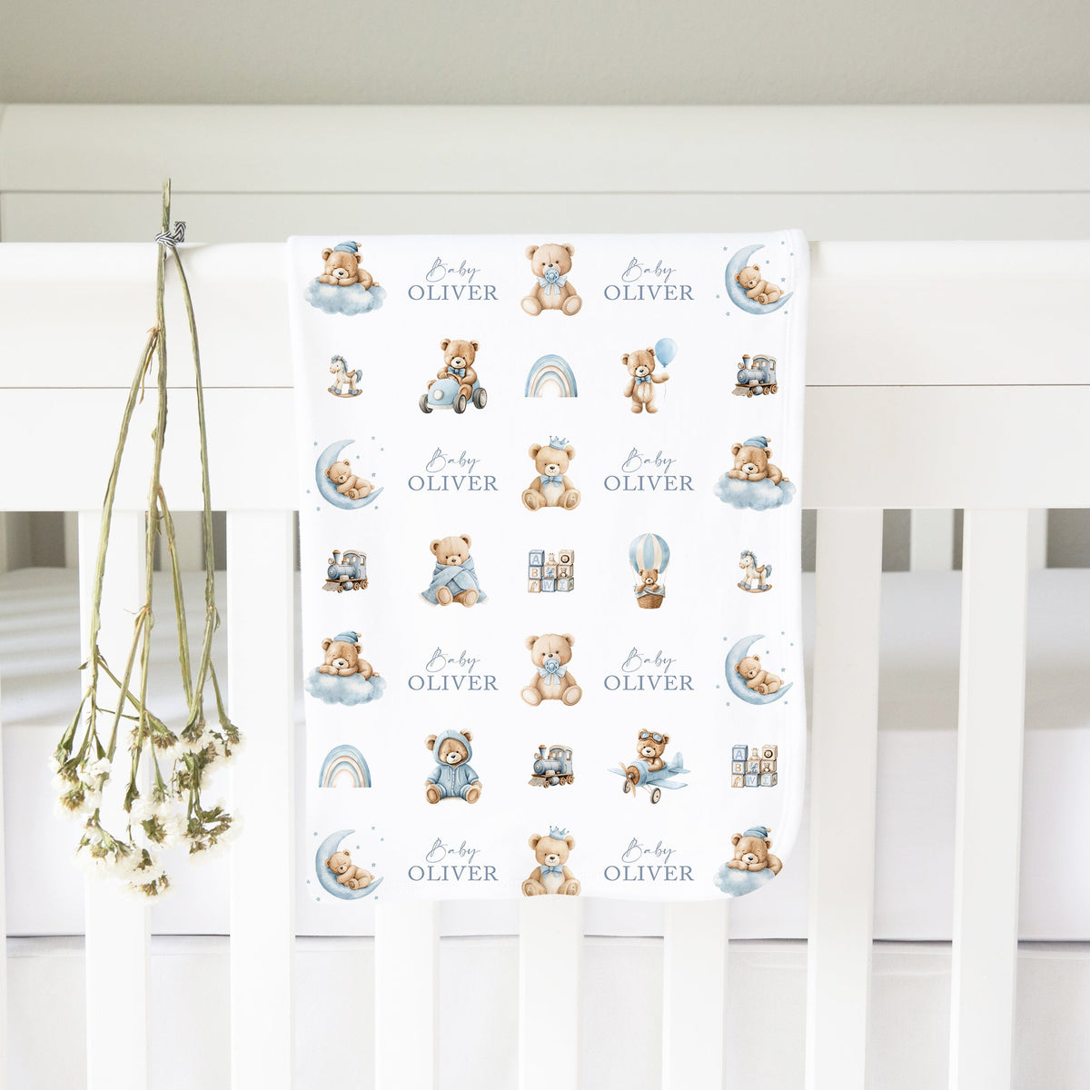 A personalised baby blanket featuring teddy bear illustrations and the name 'Baby Oliver' printed on a white crib, with dried flowers hanging from the crib rail.