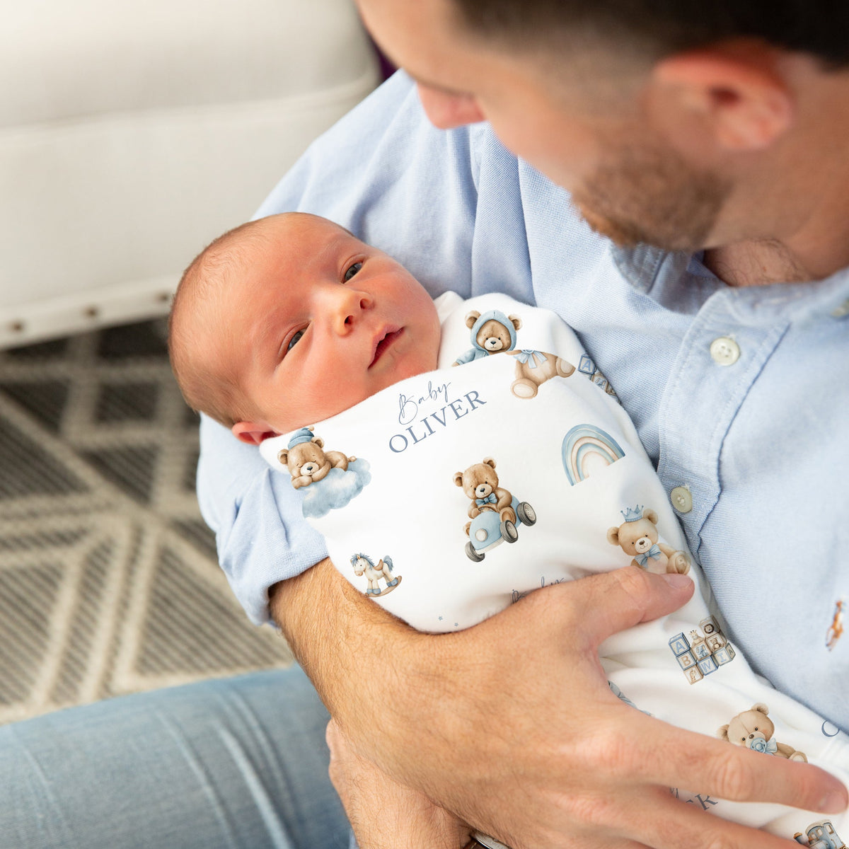 A newborn baby wrapped in a personalised white printed swaddle blanket with teddy bear illustrations and the name 'Oliver' is held by a father wearing a light blue button-up shirt. The photo is taken in a cozy indoor setting with a white sofa and patterned rug in the background.