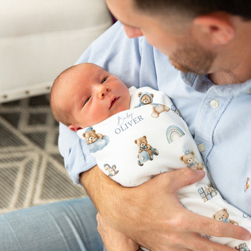 A newborn baby wrapped in a personalised white printed swaddle blanket with teddy bear illustrations and the name 'Oliver' is held by a father wearing a light blue button-up shirt. The photo is taken in a cozy indoor setting with a white sofa and patterned rug in the background.