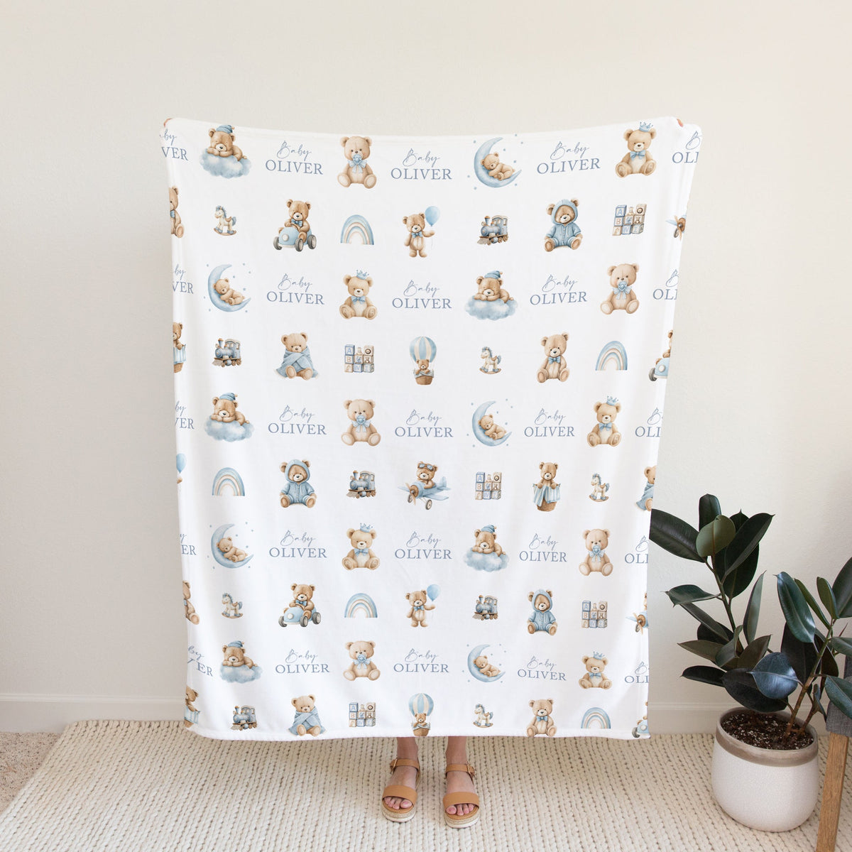 A personalised white blanket with a repeating pattern of teddy bears and the name 'Oliver' in blue, held up by a person standing in a brightly lit room
