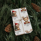 A gift box wrapped in white paper printed with teddy bear illustrations and the text 'MERRY CHRISTMAS NOAH,' tied with a twine bow, placed on green pine branches with pinecones.