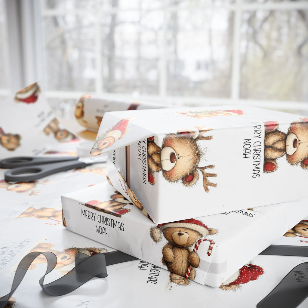 White Christmas themed wrapping paper with teddy bear illustrations and the text 'MERRY CHRISTMAS NOAH', shown wrapped around gift boxes on a table with scissors and ribbon nearby.