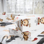 White Christmas themed wrapping paper with teddy bear illustrations and the text 'MERRY CHRISTMAS NOAH', shown wrapped around gift boxes on a table with scissors and ribbon nearby.