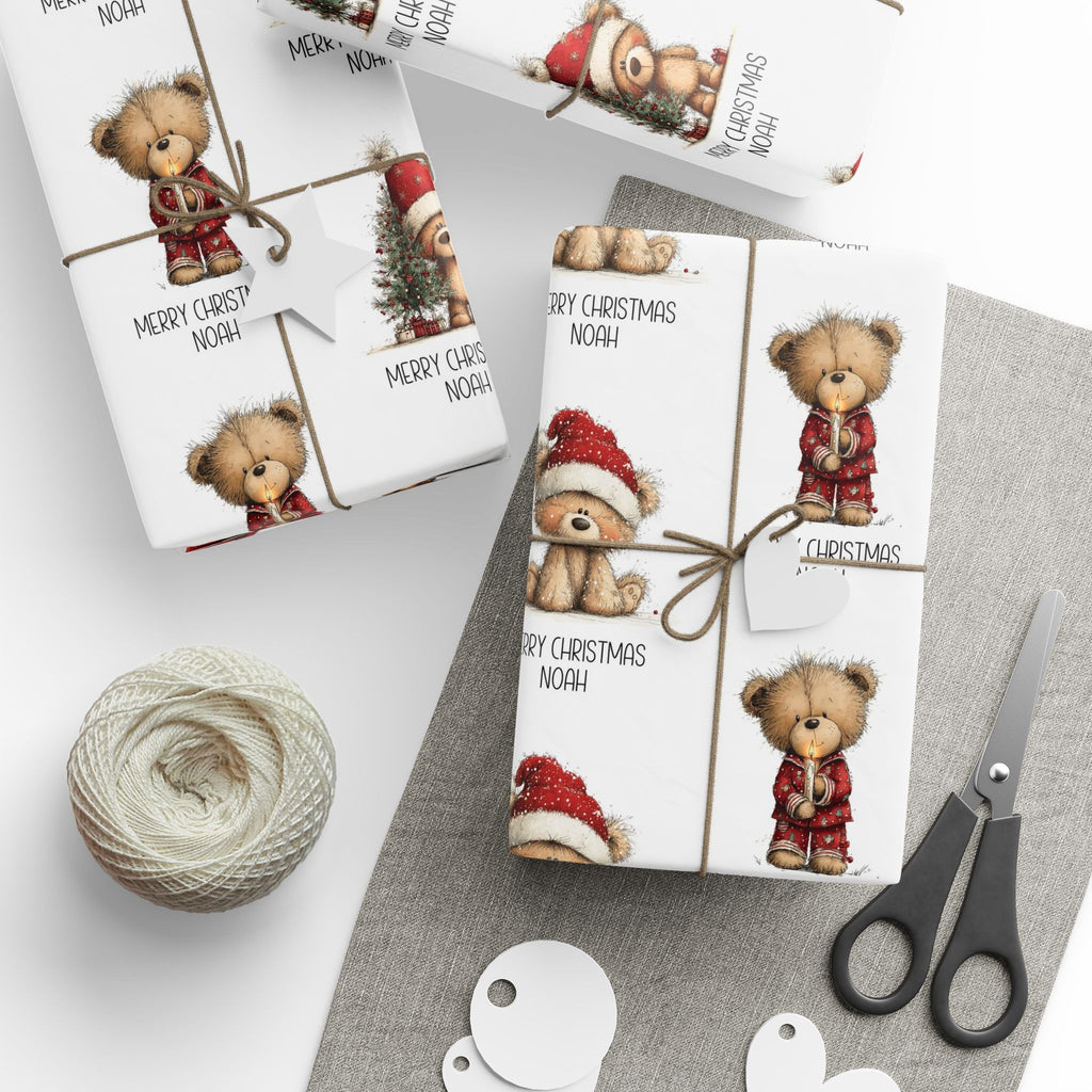 Three wrapped Christmas gift boxes with teddy bear illustrations and 'Merry Christmas Noah' text, tied with twine and styled with scissors, string, and paper tags on a grey surface.