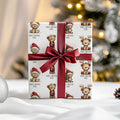 A wrapped gift box with a white paper pattern featuring teddy bears in Christmas outfits and the text 'MERRY CHRISTMAS NOAH', tied with a wide dark red satin ribbon and bow, set against a softly blurred holiday background.