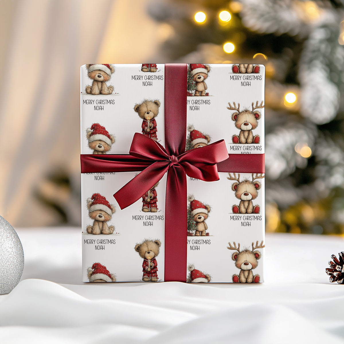 A wrapped gift box with a white paper pattern featuring teddy bears in Christmas outfits and the text 'MERRY CHRISTMAS NOAH', tied with a wide dark red satin ribbon and bow, set against a softly blurred holiday background.