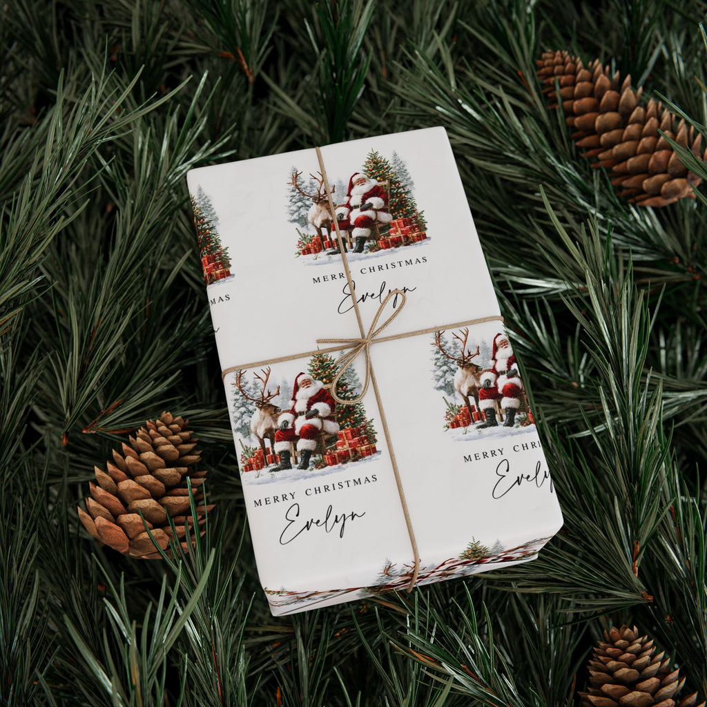 A gift box with personalised Christmas gift wrap with twine, featuring a festive holiday illustration of Santa Claus, reindeer, pine trees, and the name 'Evelyn' on a white background, styled on green pine branches with pinecones.