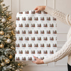A person holding a roll of wrapping paper with a repeating Santa, Christmas tree and reindeer pattern in front of a decorated holiday tree.