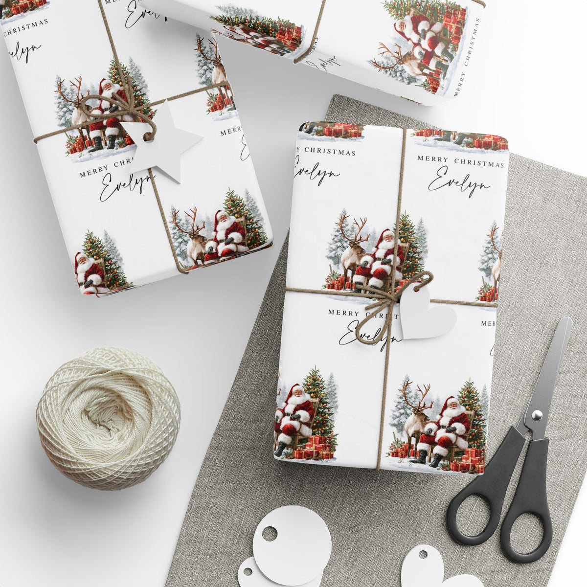 Three Christmas themed gift wraps wrapped around rectangular boxes, styled with twine, a heart-shaped tag, a ball of jute twine, scissors, and white round tags on a light grey surface.