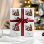A square gift box wrapped in white paper printed with four Santa and reindeer illustrations and the text 'Merry Christmas Evelyn,' tied with a wide dark red ribbon and bow, set on a white surface with a softly blurred festive background.