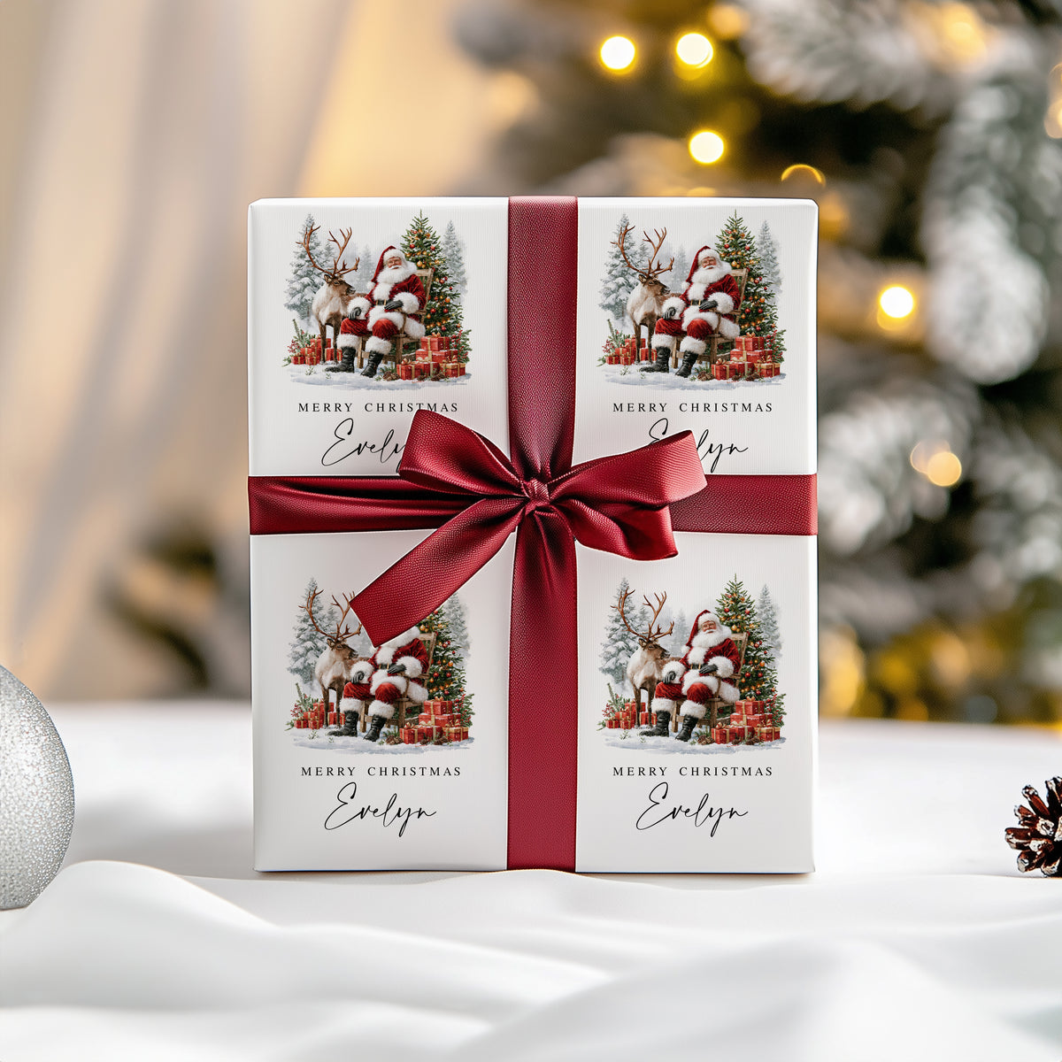 A square gift box wrapped in white paper printed with four Santa and reindeer illustrations and the text 'Merry Christmas Evelyn,' tied with a wide dark red ribbon and bow, set on a white surface with a softly blurred festive background.