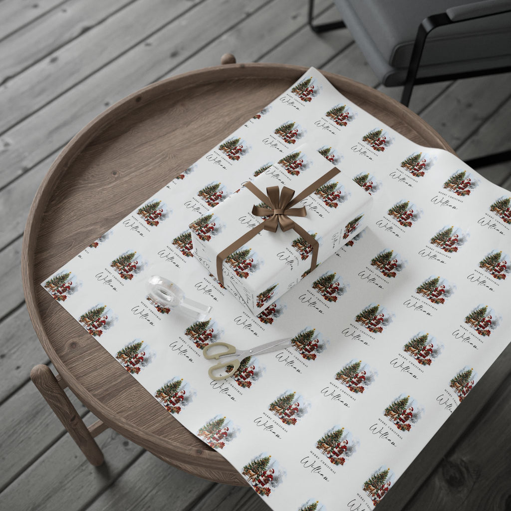 A gift box wrapped in white wrapping paper with a repeating Christmas tree, Santa Claus and Reindeer pattern with the text 'Merry Christmas William', tied with a brown ribbon, displayed on a round wooden tray over a grey deck floor.