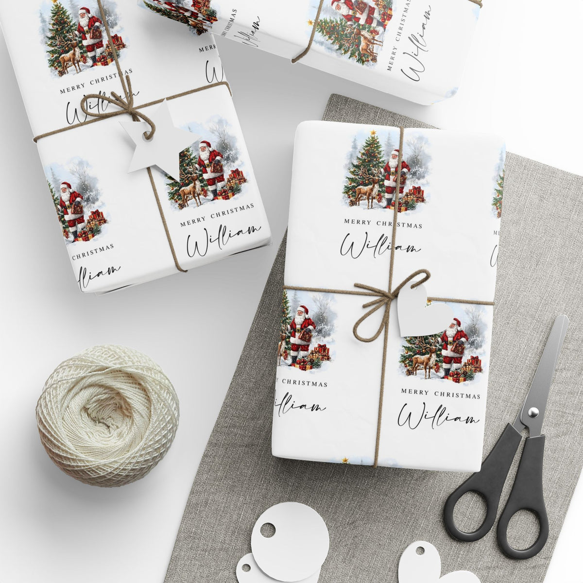 Three gift wrapped packages tied with twine on a light surface, featuring a Christmas print with Santa, trees, reindeer, and the text 'Merry Christmas William.' A ball of twine, scissors, and round tags are arranged around them.