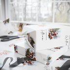White Christmas-themed gift wrapping paper and matching gift boxes with illustrations of Santa Claus, a Christmas tree, and snow, styled on a table with scissors and ribbon in front of a bright window.