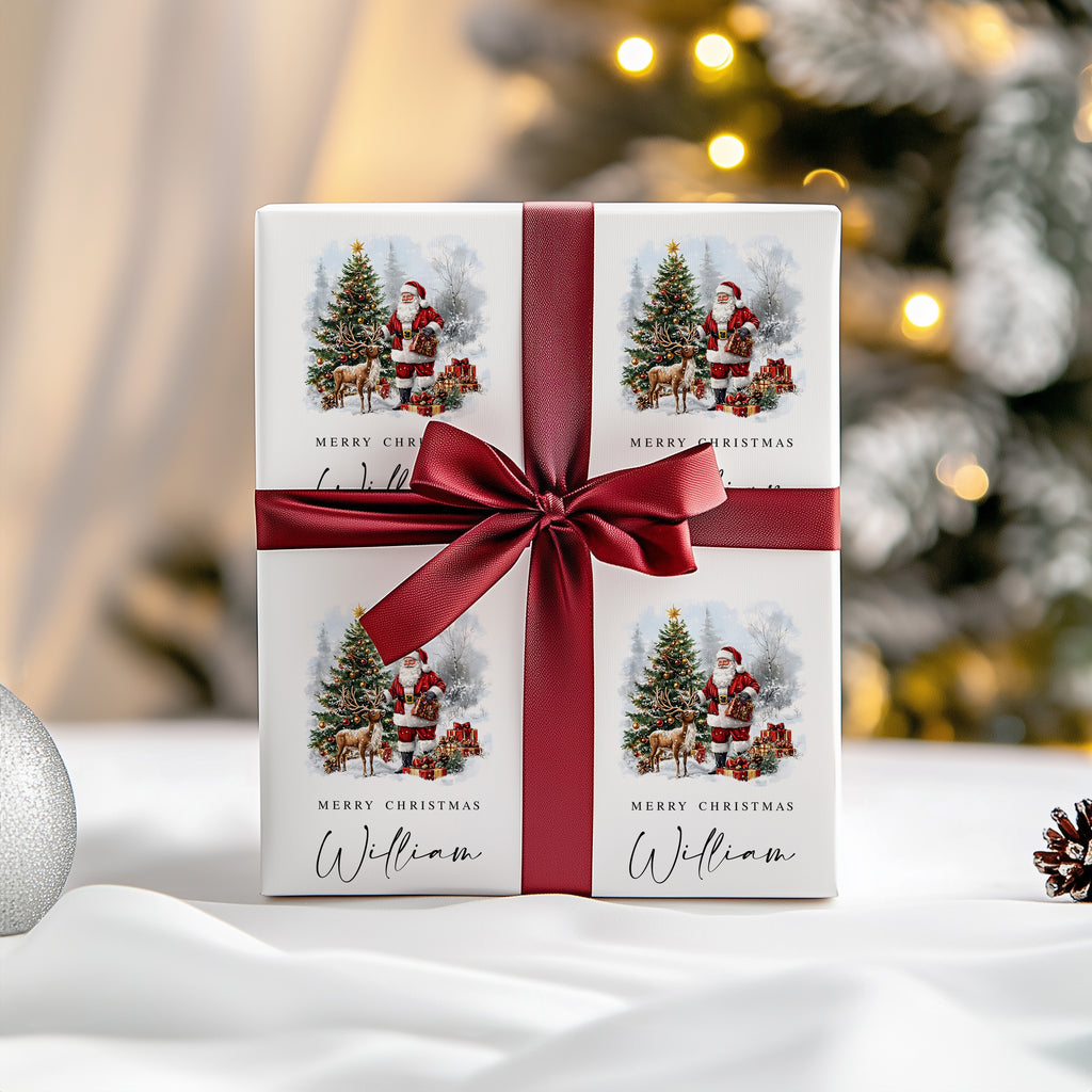 A square white gift box wrapped with a wide dark red ribbon and bow, printed with four Christmas illustrations of Santa Claus and the text 'Merry Christmas' and 'William'. The box is photographed on a white surface with a softly blurred Christmas tree and warm lights in the background.