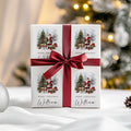 A square white gift box wrapped with a wide dark red ribbon and bow, printed with four Christmas illustrations of Santa Claus and the text 'Merry Christmas' and 'William'. The box is photographed on a white surface with a softly blurred Christmas tree and warm lights in the background.