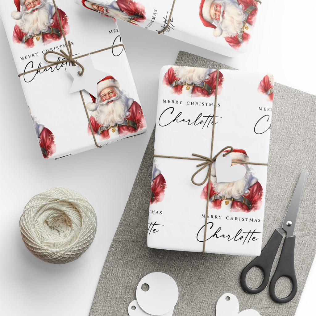 Personalised Christmas wrapping paper featuring classic watercolour Santa Claus design with the name Charlotte, wrapped gifts, twine, gift tags and scissors.