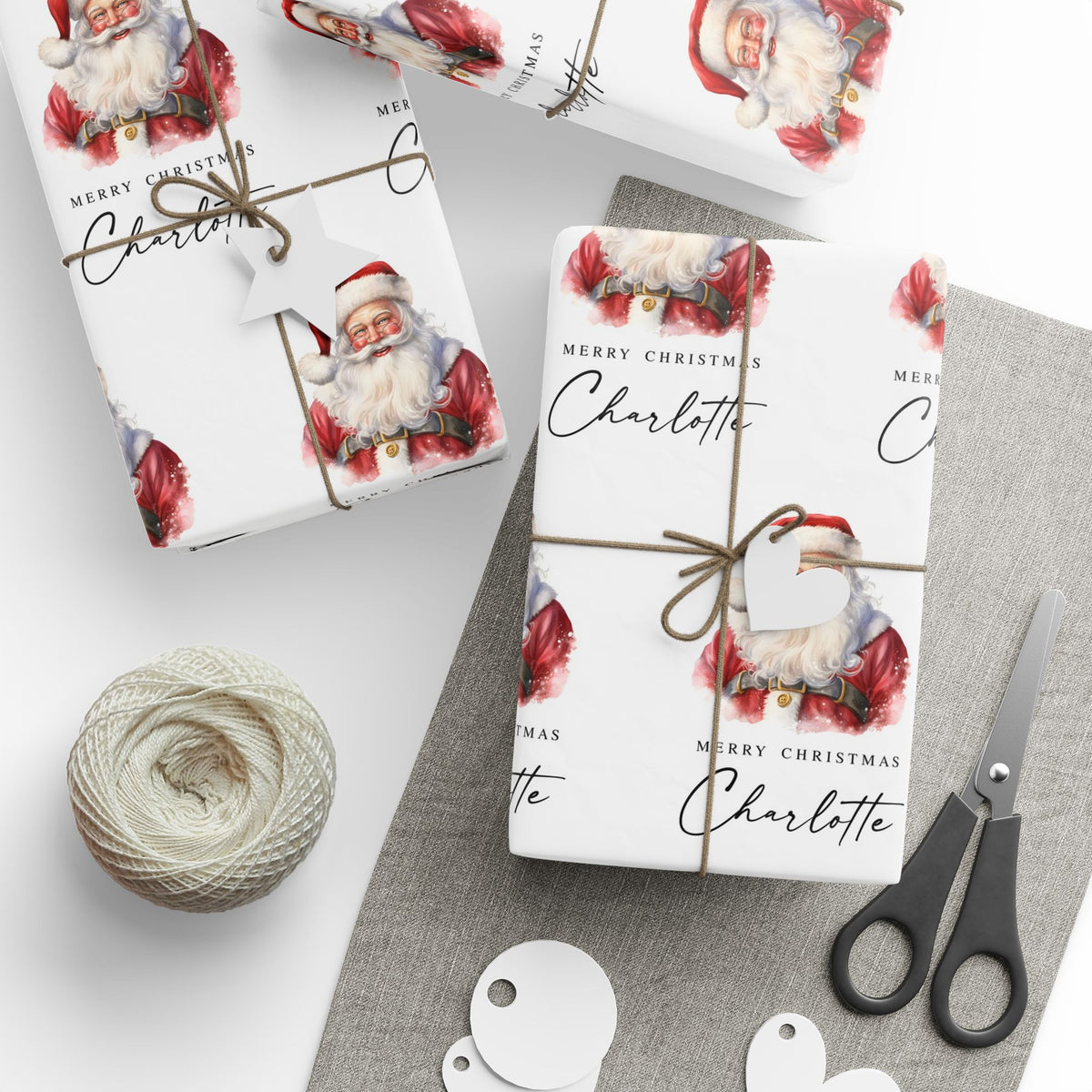 Personalised Christmas wrapping paper featuring classic watercolour Santa Claus design with the name Charlotte, wrapped gifts, twine, gift tags and scissors.