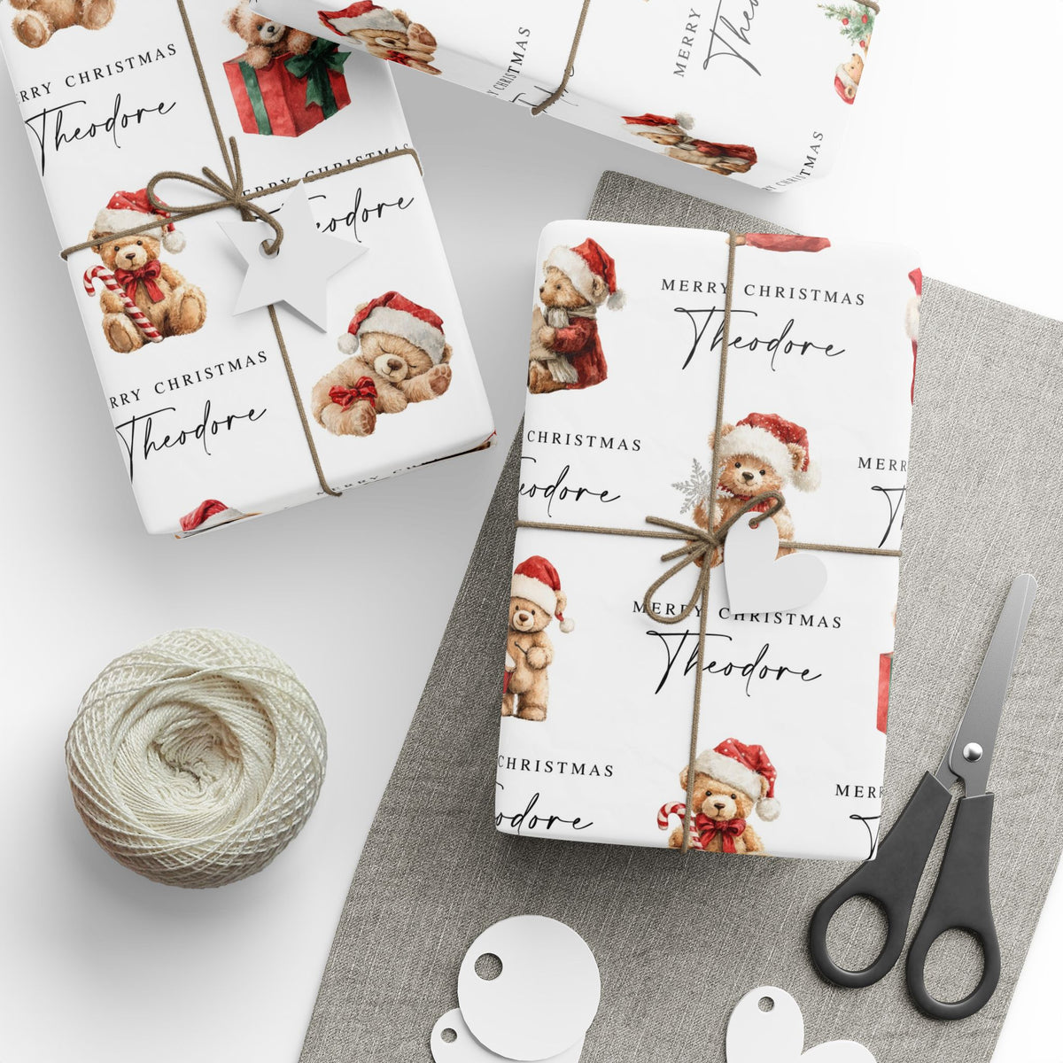Three gift wrapped boxes arranged on a light grey surface with wrapping paper printed with Christmas teddy bears, the text 'Merry Christmas Theodore.' A ball of twine, scissors, and small round tags are placed nearby as styling props.