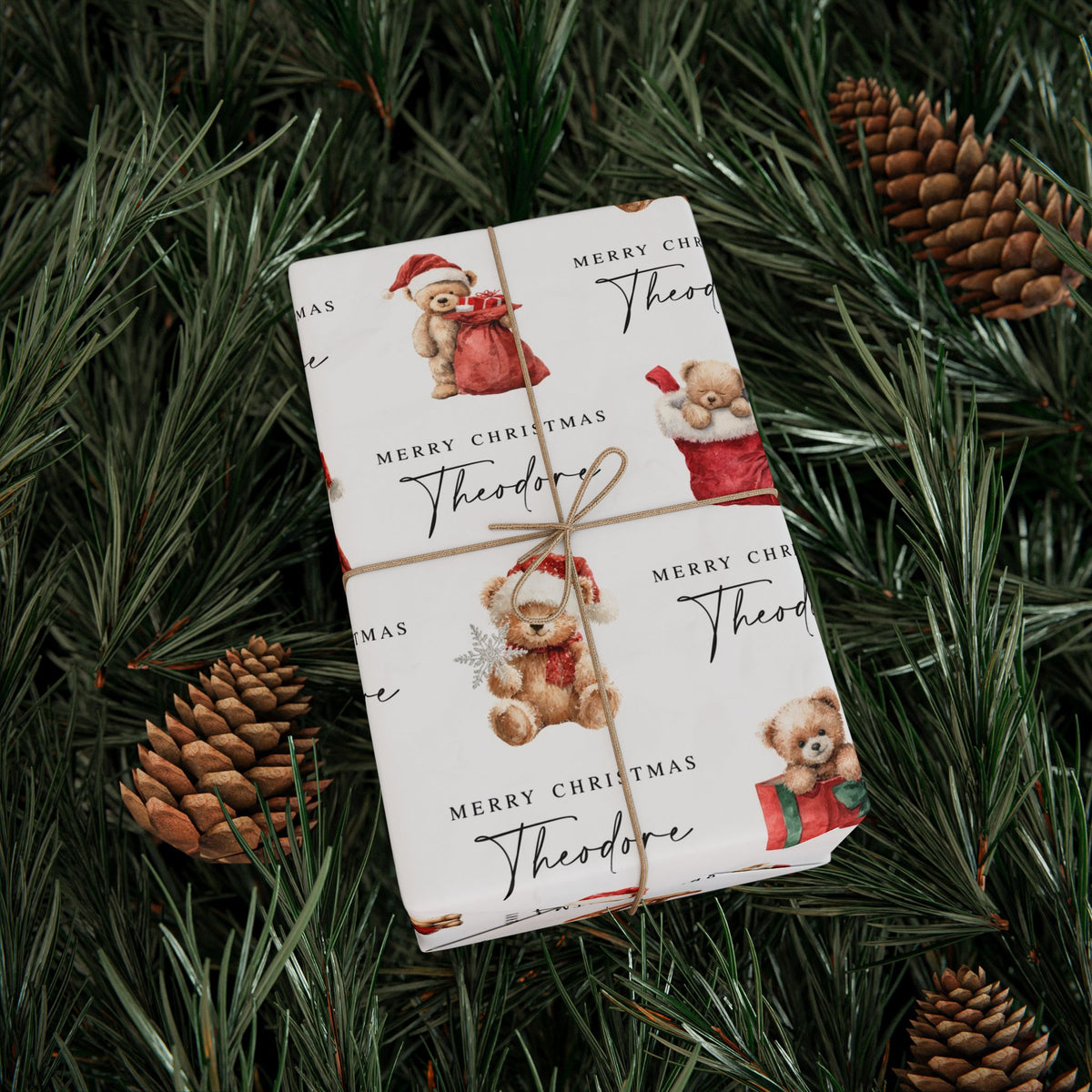 A wrapped gift box tied with twine, covered in white Christmas themed wrapping paper printed with teddy bears in Christmas outfits, and the text 'Merry Christmas Theodore,' placed on green pine branches with pinecones.