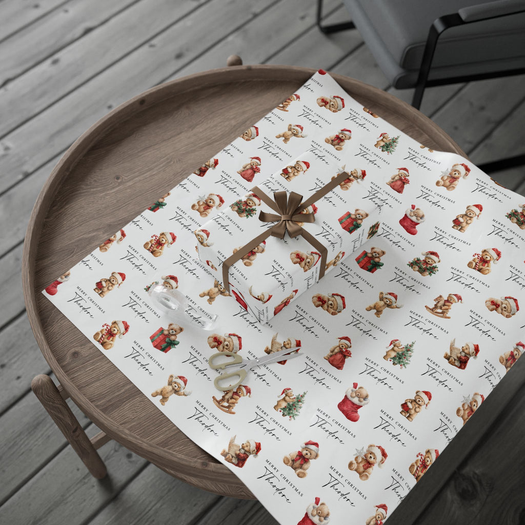 A roll of wrapping paper with a vintage teddy bear pattern, partially unrolled on a round wooden tray. The paper features a white background with a repeating print of brown Christmas teddy bears and Christmas outfits, alongside the word 'Merry Christmas' in cursive.
