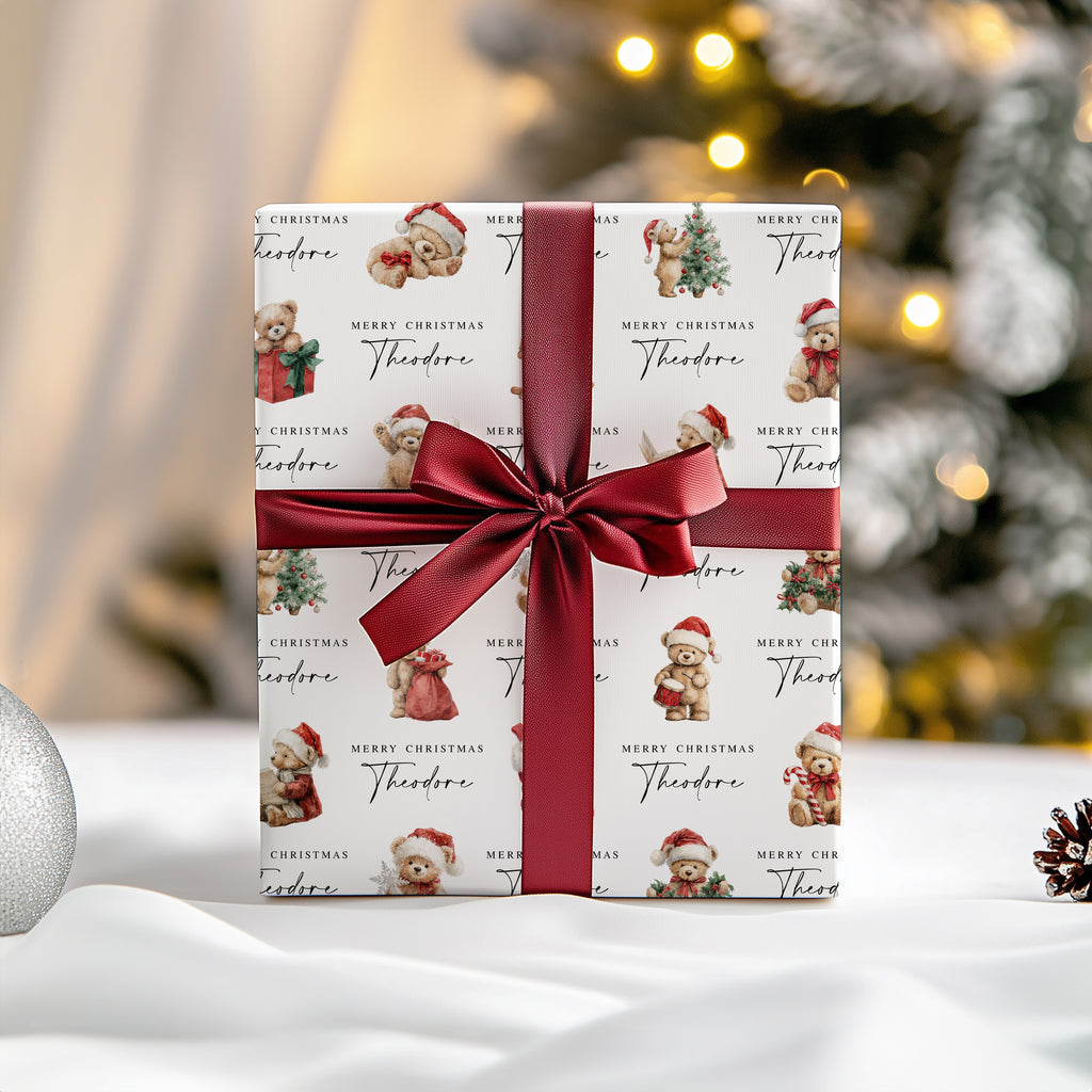A wrapped gift box with a white paper pattern featuring teddy bears in Santa hats and the text 'Merry Christmas Theodore,' tied with a wide dark red satin ribbon and bow. The scene is set on a white surface with a softly blurred Christmas tree and warm lights in the background.