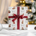 A wrapped gift box with a white paper pattern featuring teddy bears in Santa hats and the text 'Merry Christmas Theodore,' tied with a wide dark red satin ribbon and bow. The scene is set on a white surface with a softly blurred Christmas tree and warm lights in the background.