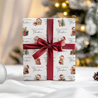 A wrapped gift box with a white paper pattern featuring teddy bears in Santa hats and the text 'Merry Christmas Theodore,' tied with a wide dark red satin ribbon and bow. The scene is set on a white surface with a softly blurred Christmas tree and warm lights in the background.