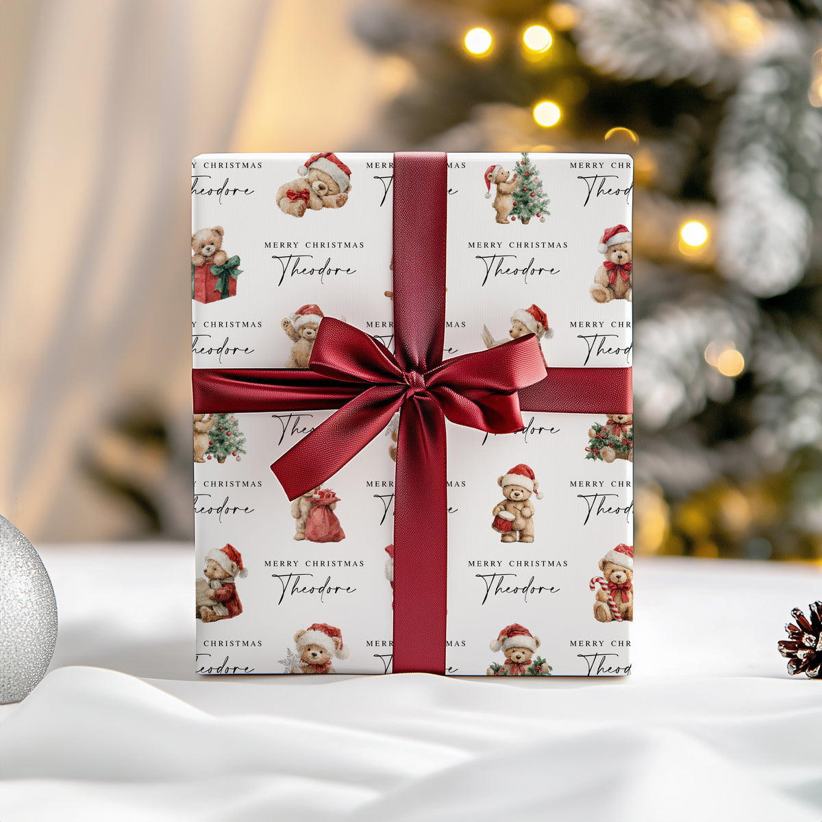 A wrapped gift box with a white paper pattern featuring teddy bears in Santa hats and the text 'Merry Christmas Theodore,' tied with a wide dark red satin ribbon and bow. The scene is set on a white surface with a softly blurred Christmas tree and warm lights in the background.