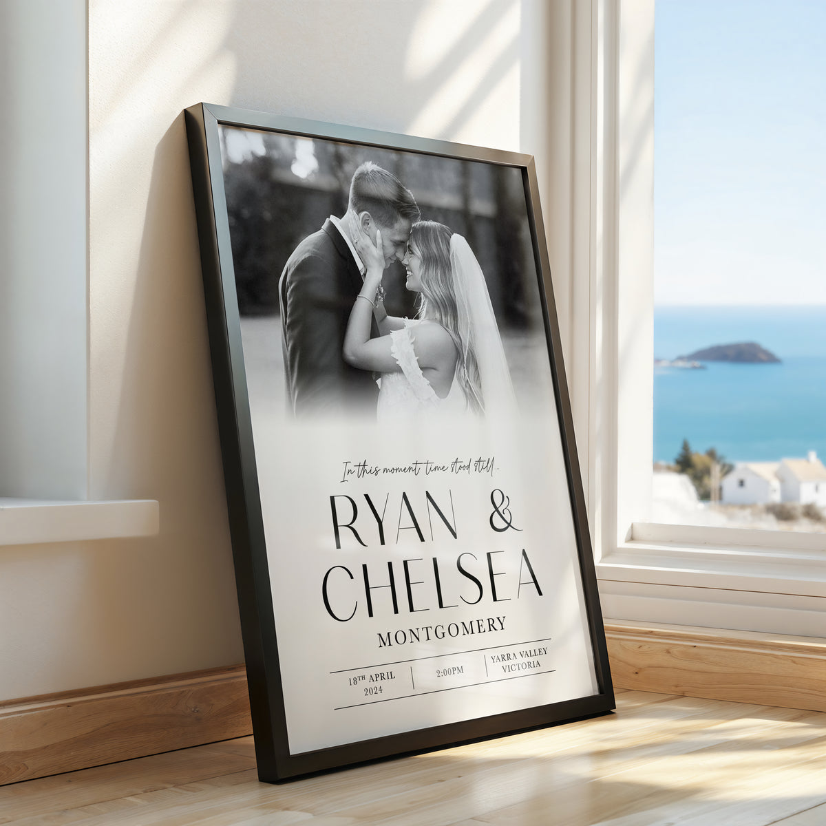A framed custom photo print leaning against a wall next to a window, showing a black and white wedding photograph of a couple with the names 'Ryan & Chelsea' and the date '18th April 2024' printed on it. The room has light wood floors and a bright blue ocean view outside the window.