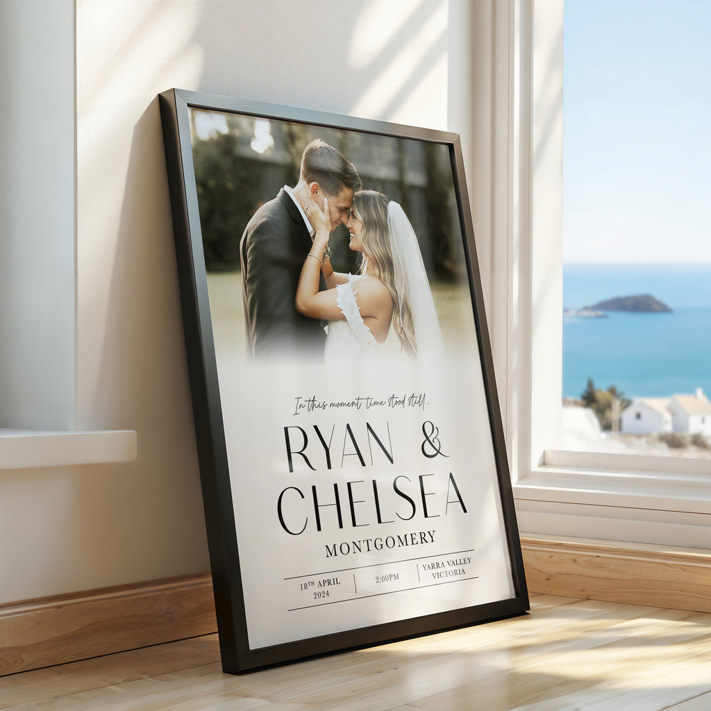 A framed custom photo print leaning against a wall next to a window, showing a wedding photograph of a couple with the names 'Ryan & Chelsea' and the date '18th April 2024' printed on it. The room has light wood floors and a bright blue ocean view outside the window.