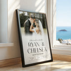A framed custom photo print leaning against a wall next to a window, showing a wedding photograph of a couple with the names 'Ryan & Chelsea' and the date '18th April 2024' printed on it. The room has light wood floors and a bright blue ocean view outside the window.