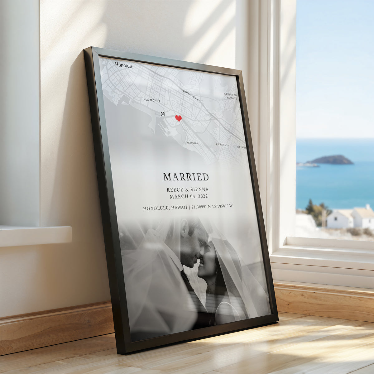 A framed custom map wedding print leaning against a window on a wooden floor, showing a black-and-white map with a small red heart, the word 'MARRIED,' couple names and date, and a soft couple portrait at the bottom. A real interior setting and ocean view are visible outside the window.