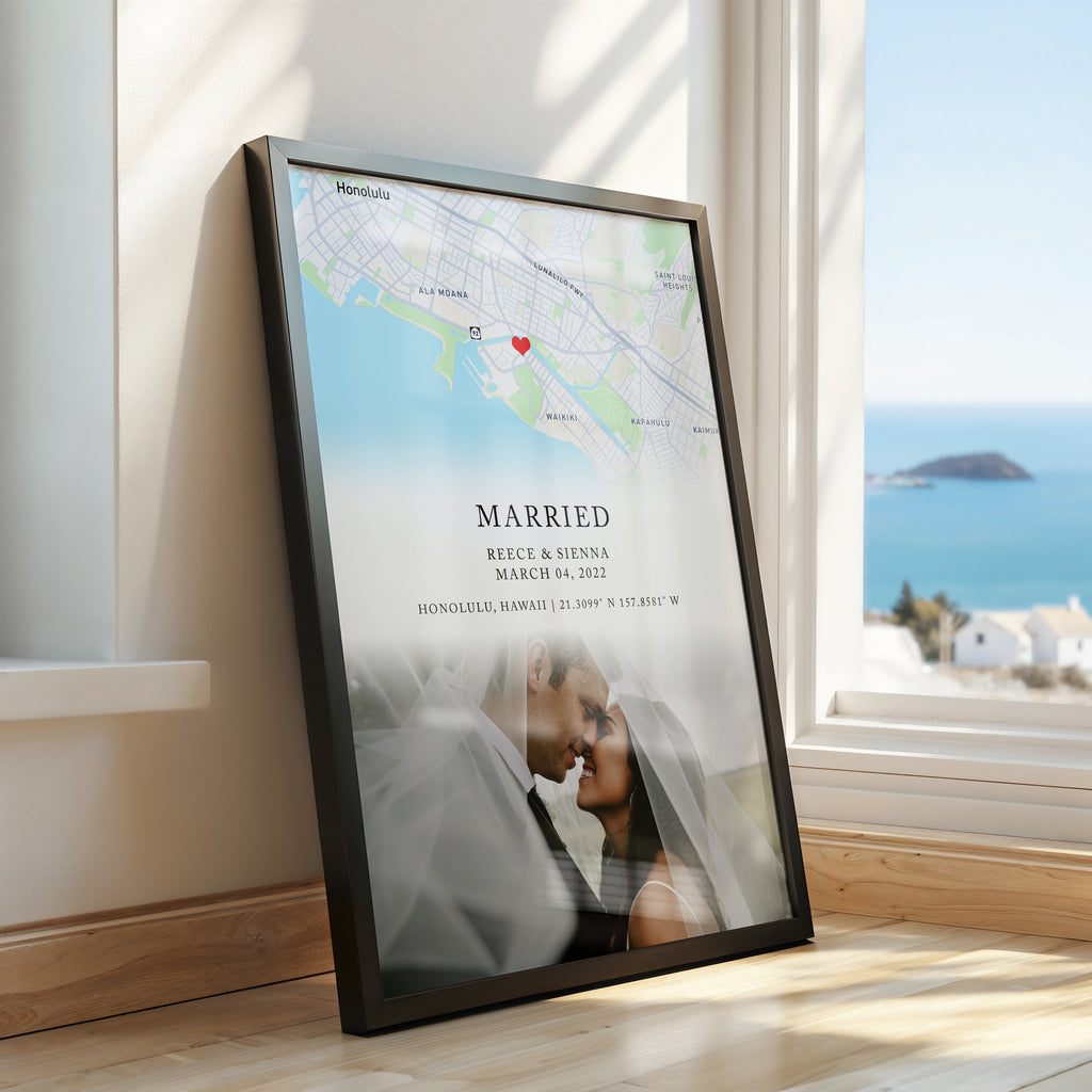A framed custom map and photo print leaning against a window on a wooden floor, with a coastal outdoor view visible outside. The print includes a map of Honolulu, a red heart marker, the word 'MARRIED,' names and date of a couple, and a faded photo of the couple at the bottom.