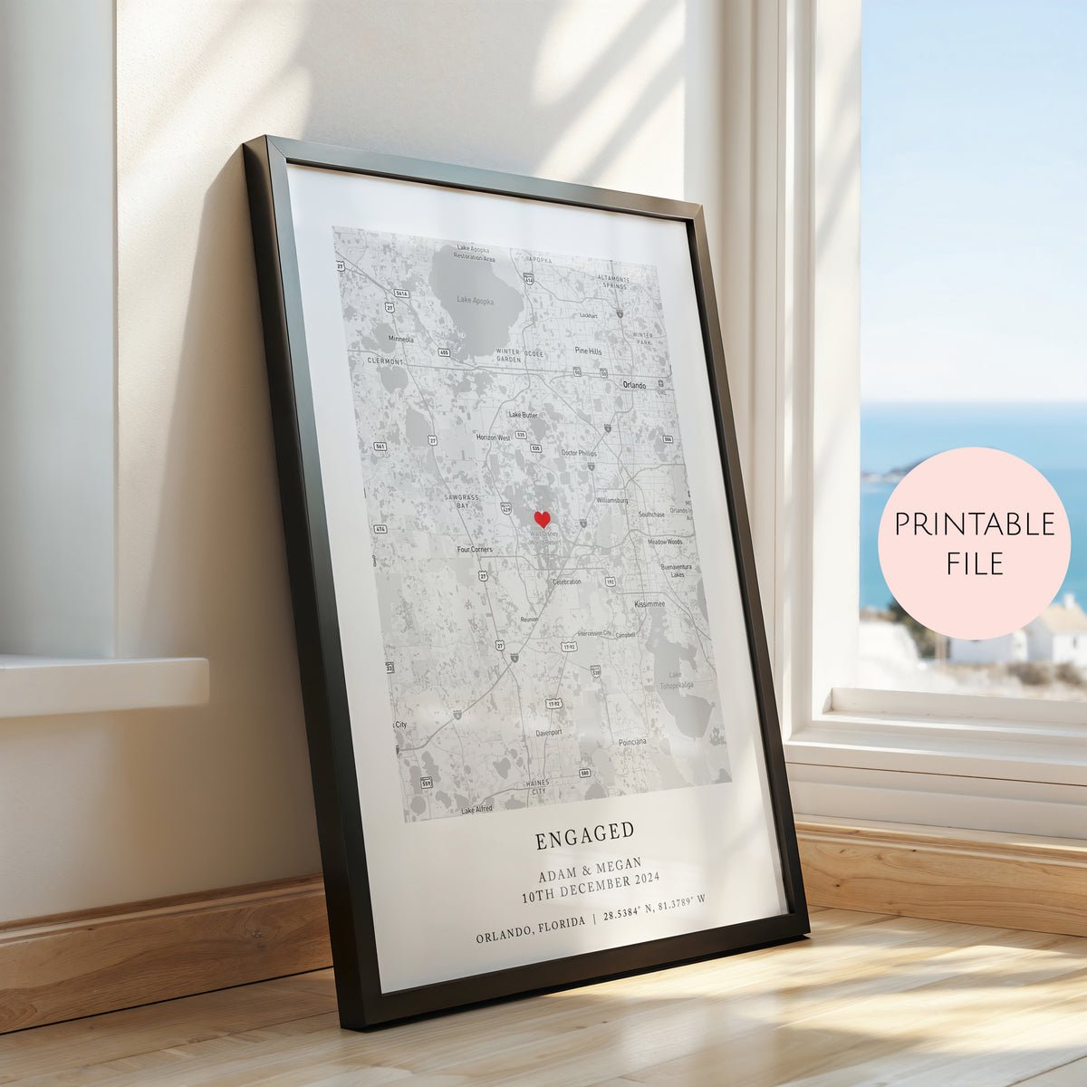 A framed personalized map print leaning against a window on a wooden floor, with a pink circular badge reading 'PRINTABLE FILE' on the right. The print shows a black and white city map with a small red heart marker and the word 'ENGAGED' at the bottom.