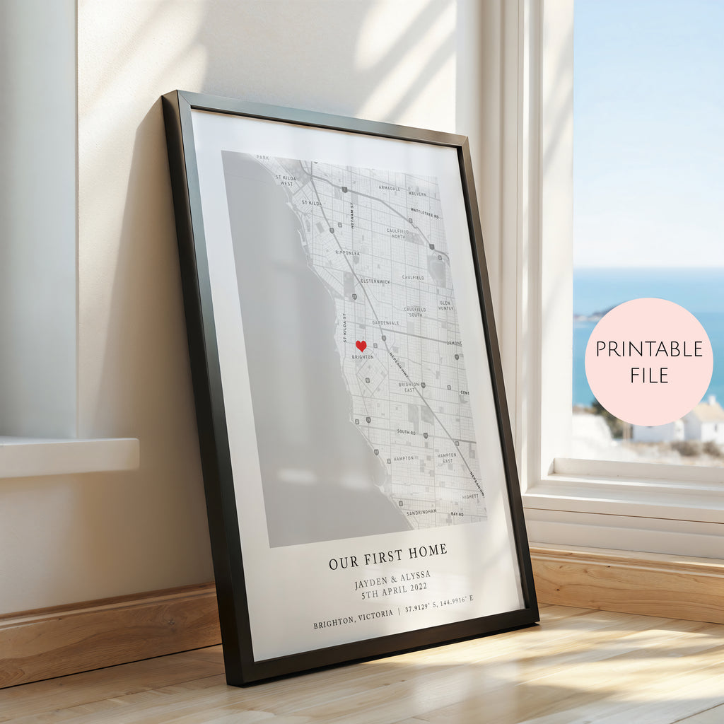 A framed personalised map print leaning against a window on a wooden floor. The print shows a street map of Brighton, Victoria with a small red heart marking a location, plus the text 'OUR FIRST HOME' and custom names/date.