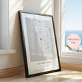 A framed personalised map print leaning against a window on a wooden floor. The print shows a street map of Brighton, Victoria with a small red heart marking a location, plus the text 'OUR FIRST HOME' and custom names/date.