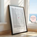 A framed personalised map print leaning against a window on a wooden floor. The print shows a street map of Brighton, Victoria with a small red heart marking a location, plus the text 'OUR FIRST HOME' and custom names/date.